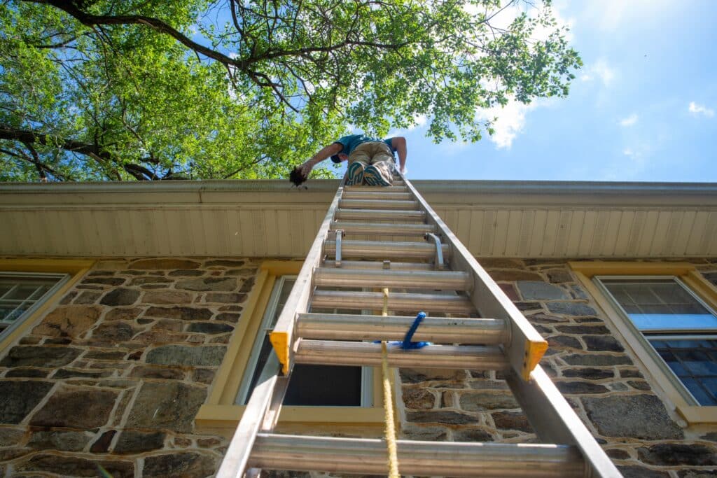 Low angle view of man on ladder cleaning gutters of stone home